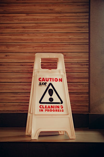 A white plastic 'Caution: Cleaning in Progress' floor-standing sign with a black triangular warning symbol and red text, placed on a hardwood floor next to a wooden paneled wall. The sign indicates on