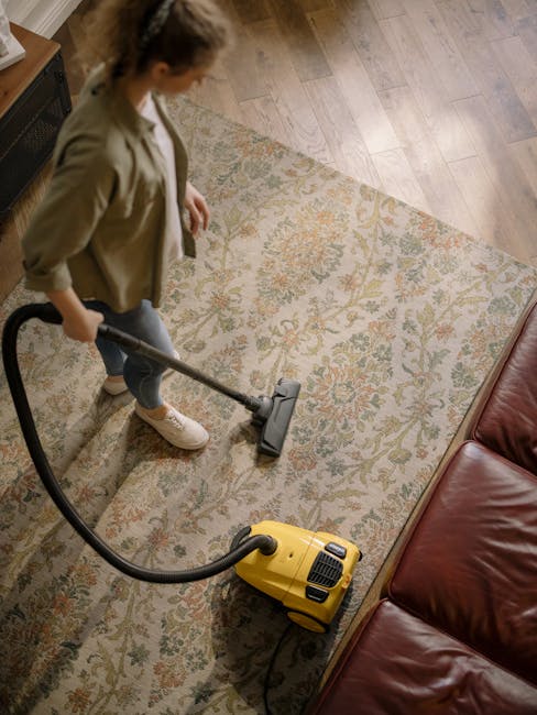 Two professional cleaning technicians from Cleaners Belgravia, wearing beige uniforms, standing back-to-back in a well-lit bedroom. One holds a vacuum cleaner, and the other is holding a stream mop. T