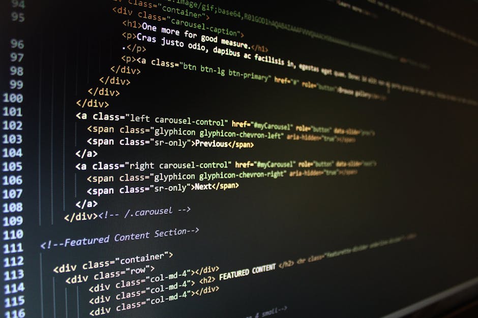 Close-up of a person's hand operating a silver laptop with a black keyboard on a wooden desk. The screen displays lines of colorful code in a text editor, suggesting software or web development. The p